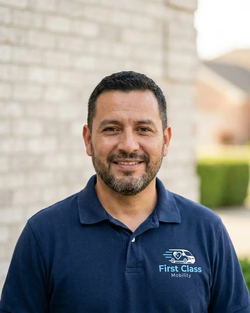 A First Class Mobility driver in a navy polo shirt with the company logo smiles in front of a light brick wall.